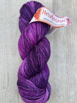 Ein handgefärbter Strang Wolle in leuchtenden Purpur- und Magentatönen mit dunklen violetten Akzenten. Das Garn liegt locker gedreht auf hellem Holz, das Etikett zeigt das Herzfasern-Logo.